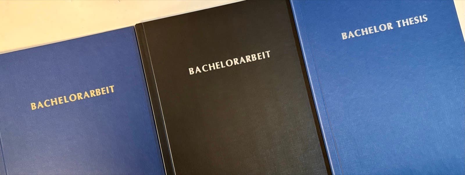 Drei geprägte Hardcover aus unserem Copyshop: Blau mit Gold-, Schwarz und Blau mit Silberprägung – „Bachelorarbeit“ und „Bachelor Thesis“