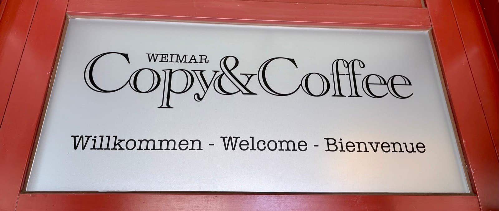 Türschild von Copy&Coffee Weimar: „Willkommen – Welcome – Bienvenue“ auf der roten Ladentür